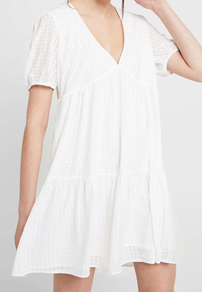 Femme portant une robe babydoll blanche à manches courtes, col en V, à volants avec une texture subtile à carreaux, posant avec un bras levé.