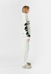 Sudadera blanca de gran tamaño con texto y acentos negros, combinada con pantalones blancos a juego. Presenta puños acanalados y calzado texturizado beige.