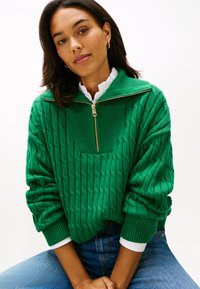 Zöld kötött pulóver fél cipzáras nyakrésszel, texturált mintával és bordázott szegélyekkel. Fehér ing gallérja alatt viselve.