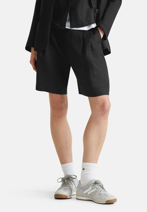 Person, der schwarze knielange Shorts, eine schwarze Jacke, weiße Socken und graue Turnschuhe trägt, steht mit einer Hand in der Tasche.