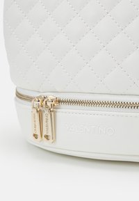Vit quiltad handväska med guldiga detaljer och dubbla dragkedjor. Har ett präglat varumärkeslogotyp och en slät texturerad botten.