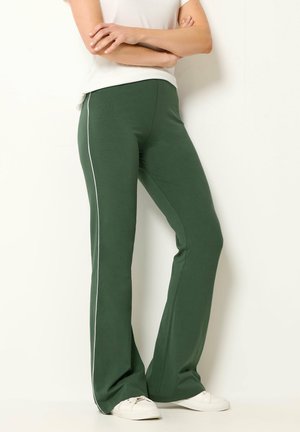 Person trägt grüne ausgestellte Hose mit weißem Seitenstreifen, weiße Sneakers und weißes kurzärmeliges Oberteil, steht mit verschränkten Armen vor einer weißen Wand.
