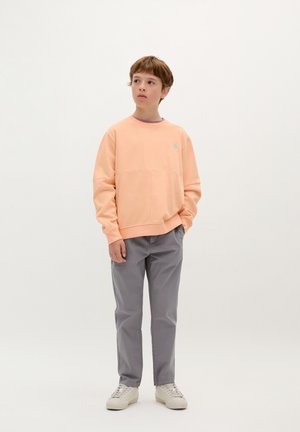 Felpa color pesco chiaro con frontale texturizzato e piccolo logo teal, abbinata a pantaloni grigi e sneakers avorio, su uno sfondo neutro.
