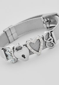 Bracelet en maille argentée ornée de charmes en forme de cœur et de cadenas, embellie de strass et de pointes, offrant un design texturé et superposé.