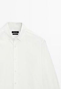 Chemise blanche à col classique, avec une texture de tissu lisse et une coupe ajustée. Étiquette visible sur le col.