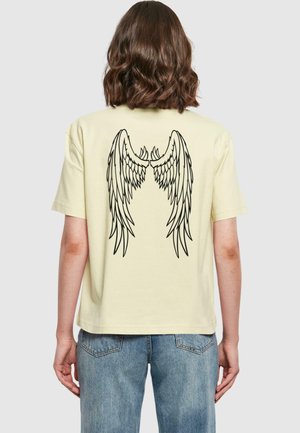 Person mit schulterlangem braunem Haar, der ein cremefarbenes T-Shirt mit schwarzem Engel-Flügel-Design auf dem Rücken und blaue Jeans trägt.