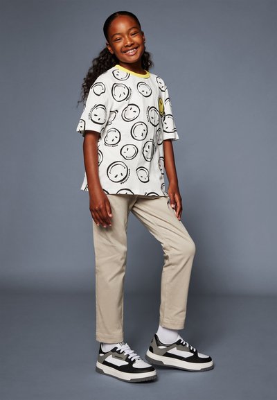 Maglietta bianca con un grande motivo di faccina sorridente nera e collo giallo, abbinata a pantaloni beige e sneakers nere con accenti bianchi.