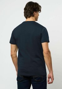 Vyras stovintis nugara, dėvintis paprastą tamsiai mėlyną trumpomis rankovėmis marškinėlį ir tamsius džinsus prieš paprastą šviesų foną.