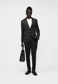 Άνδρας φορώντας σκούρο καφέ κοστούμι με λευκό t-shirt και μαύρα παπούτσια, κρατώντας μαύρη δερμάτινη τσάντα, στέκεται μπροστά σε απλό ανοιχτό φόντο.