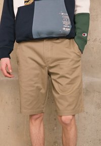 Shorts beiges avec une texture lisse, dotés de poches latérales et d'une coupe droite. Associés à un sweat-shirt multicolore bleu et vert.