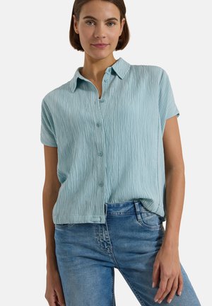 Donna che indossa una camicia azzurra a maniche corte texturizzata abbottonata e infilata in jeans blu medi, in posa con una mano lungo il fianco.