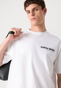 Balta kokvilnas T-kreklā ar īsām piedurknēm, kas ir rotāta ar melnu logotipu kreisajā pusē. Modelis tur melnu ādas somu.