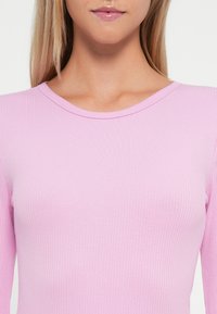 Haut à manches longues rose côtelé avec un col rond. Le tissu a une texture lisse et un design ajusté, mettant en avant son extensibilité.