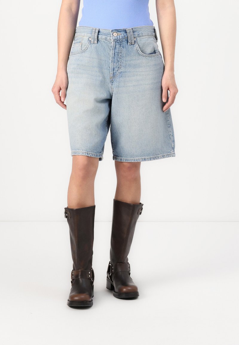 Svetlo modri denim kratki hlače s klasičnim krojem, z gumbnim pasom, štirimi žepi in zvitimi robovi, v kombinaciji s svetlomodrimi čevlji do kolen.