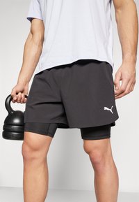 Pantalones cortos deportivos negros con un interior de malla, que presentan un logo blanco de Puma. Combinados con una camiseta gris clara, la persona sostiene una kettlebell negra.