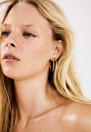 Femeie blondă cu pistrui deschiși, purtând cercei aurii rotunzi, privind ușor în sus și într-o parte pe un fundal alb simplu.