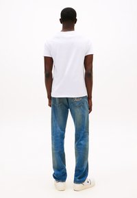 Man som står vänd bortåt, klädd i en enkel vit t-shirt, blå jeans med "Tommy Jeans"-broderi och vita sneakers.