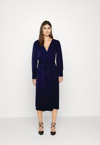 Vestido envolvente azul-marinho com mangas longas, decote em V, laço na cintura e comprimento midi. Usado com sapatos de salto preto com biqueira pontiaguda e tiras cruzadas no tornozelo.