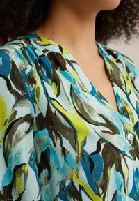 Närbild av en person som bär en V-ringad blus med ett färgglatt abstrakt lövmönster i blå, grön och brun nyanser.