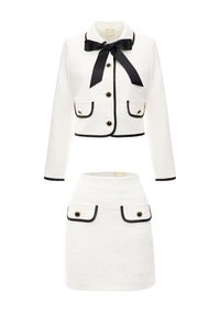 Biała żakiet z tweedu z czarnymi wykończeniami, długimi rękawami, muszką i złotymi guzikami; zestawiony z pasującą białą spódnicą z tweedu z kieszeniami.