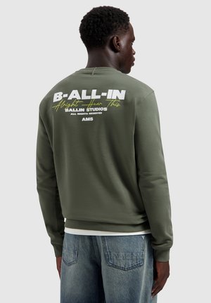 Man met een olijfgroene trui met witte en gele tekst "B-ALL-IN Alright Hear This BALLIN STUDIOS AMS" op de achterkant.
