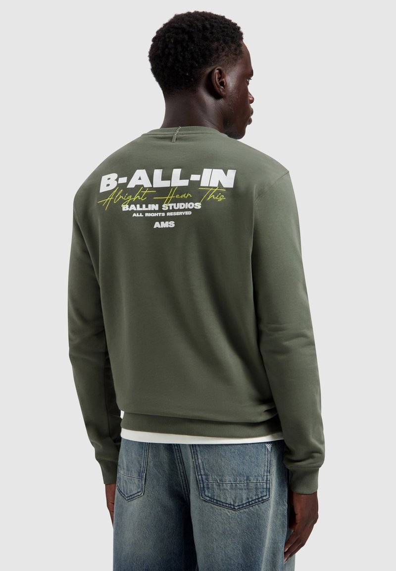 Man met een olijfgroene trui met witte en gele tekst "B-ALL-IN Alright Hear This BALLIN STUDIOS AMS" op de achterkant.