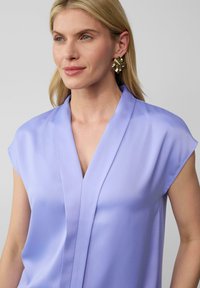 Lichtpaarse blouse met korte mouwen en een V-hals ontwerp, met een gladde, zachte textuur en minimalistische details. Gouden abstracte oorbellen.
