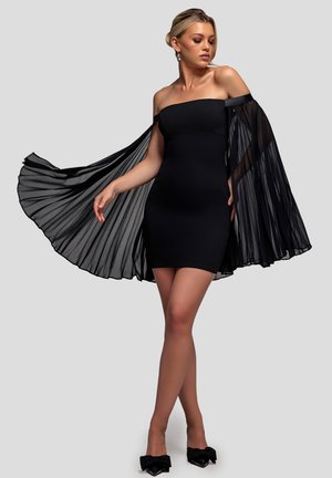 Mujer posando con un vestido mini negro con hombros descubiertos, mangas capa plisadas transparentes y tacones negros de punta afilada con lazos.