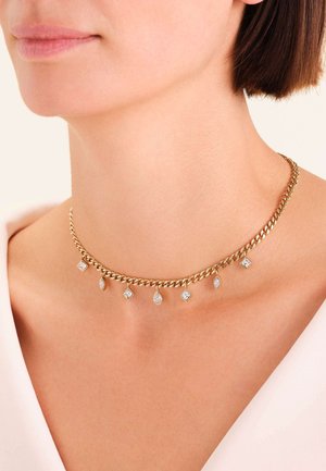 Collana di catena d'oro con charm a forma di goccia e quadrato in pietra trasparente alternati, indossata da una donna con una maglietta bianca.