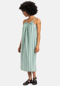 Světle zelené a šedé pruhované bezramínkové šaty se skládacím výstřihem, střední délky, vyrobené z měkké tkaniny, nošené s černými sandály.