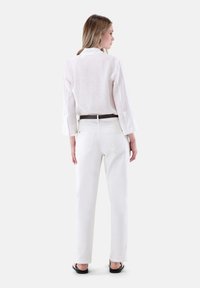 White linen shirt with a back pleat, paired with high-waisted white jeans. Brown belt, relaxed fit. Black sandals complete the outfit.