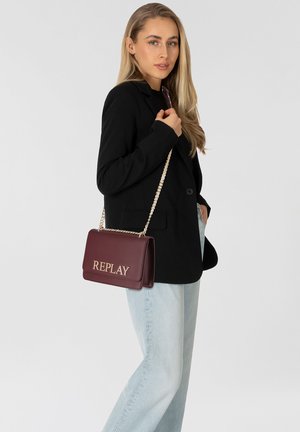 Burgunderfarbenes Leder-Crossbody-Tasche mit goldfarbener "REPLAY"-Logo, Kettenriemen, rechteckiger Form und glatter Textur. Getragen mit einem schwarzen Blazer und hellblauen Jeans.