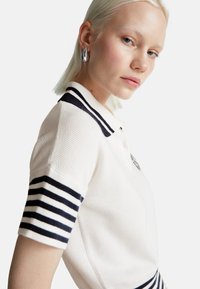 Blondýnka s krátkými vlasy, oblečená v bílé pletené polokošili s černými pruhy a stříbrnými kapkovými náušnicemi, dívá se na kameru.