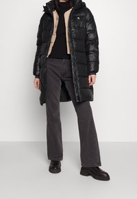 Czarna kurtka puchowa z kapturem, charakteryzująca się błyszczącym wykończeniem i pikowanym wzorem, noszona na beżowej kamizelce i czarnym swetrze, zestawiona z ciemnymi d�żinsami.