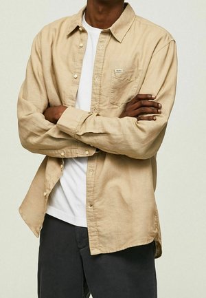 Homme à la peau foncée portant une chemise beige à boutons par-dessus un t-shirt blanc, bras croisés, et pantalon foncé devant un fond clair uni.