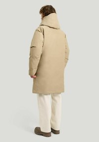 Beige lång parka med huva, som har en slät yta och en avslappnad passform. Kombinerad med krämfärgade byxor och bruna skor.
