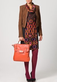 veste en daim marron sur une robe sombre à motifs circulaires orange et violet, accompagnée d'un sac en cuir orange et de collants bordeaux.