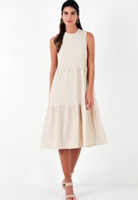 Beige sleeveless dress with a high neckline, tiered design, and textured fabric. The hemline falls below the knee, paired with neutral shoes.