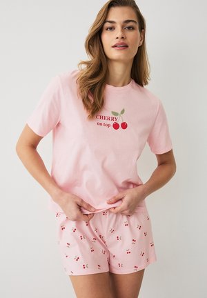 Kvinde iført lyserødt pyjamasæt med kirsebærmotiver, T-shirt med teksten "CHERRY on top" og matchende shorts med kirsebærprint.