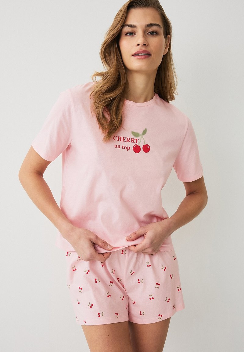 Vrouw draagt een roze pyjamaset met kersenmotieven, een T-shirt met de tekst "CHERRY on top" en bijpassende shorts met kersenprint.