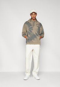 Hoodie tie-dye marron avec une poche avant, porté avec un pantalon crème et des baskets blanches ; comprend un petit motif sur la poitrine et la capuche.