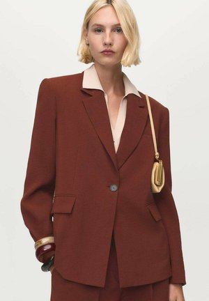 Blonde woman wearing a rust-colored tailored blazer over a beige shirt, carrying a small beige shoulder bag and chunky bracelets.