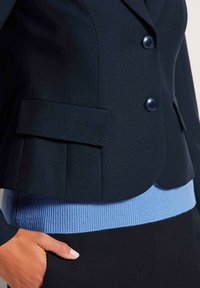 Marineblazer met twee knopen, een getailleerd ontwerp, gedragen over een lichtblauwe geribde tailleband. Heeft een zijzak en een gestructureerde stof.