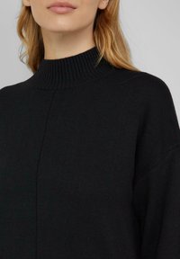 Schwarzer gestrickter Pullover mit hohem, geripptem Kragen, lockerem Sitz und strukturiertem Muster, der einen weichen Stoff und klare Linien präsentiert.