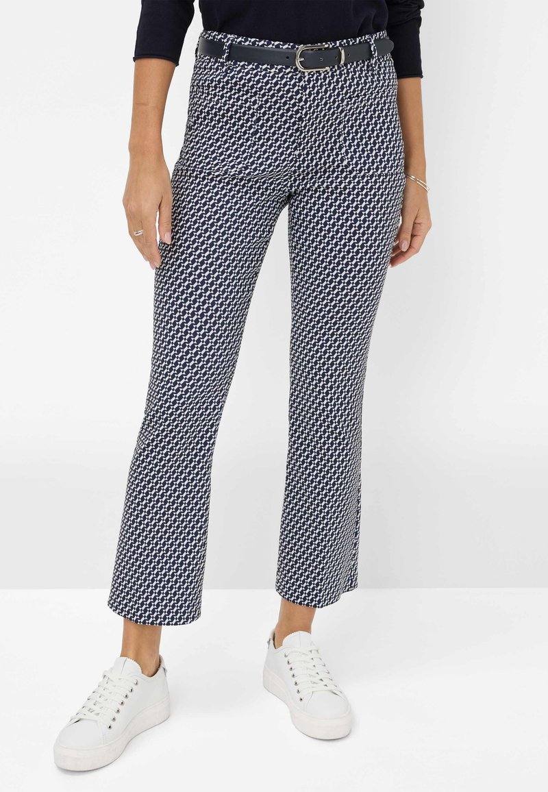 Femme portant un pantalon court à motifs bleu marine et blanc avec une ceinture noire et des baskets blanches, debout devant un fond uni.