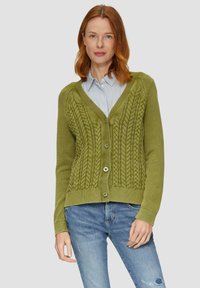 Zielony, dziergany kardigan z głębokim dekoltem w kształcie V, złożonym z misternych wzorów w warkocze i guzikami z przodu. Noszony na niebieską i białą koszulę w paski.