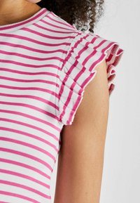 Różowo-biały top w paski z prążkowaną teksturą, z falbankowymi rękawami. Zbliżenie podkreśla wzór na ramieniu i rękawie.