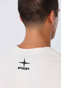 Camiseta crema con un diseño bordado en negro que incluye el texto "PXP" y un símbolo de estrella en la parte posterior. Mangas cortas y escote redondo.