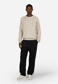 Μπεζ πουλόβέρ με πλέξη κασμίρ και στρογγυλή λαιμόκοψη, συνδυασμένο με μαύρο κομψό παντελόνι και λευκά αθλητικά παπούτσια με υφή και διακριτικό λογότυπο.