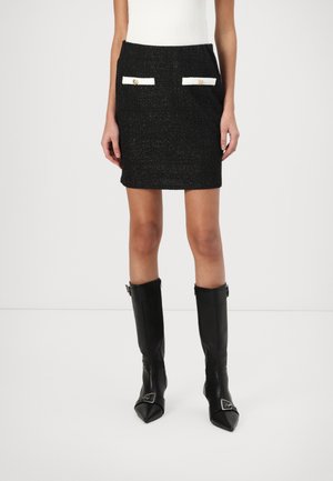 Jupe mini en tweed noir avec une finition texturée, dotée de deux poches blanches à rabat avec des boutons dorés. Assortie à des bottes en cuir noir montantes.